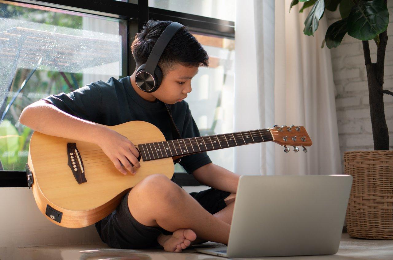 Jeune guitariste concentré utilisant une application d’apprentissage avec un professeur en arrière-plan, illustrant l'apprentissage en ligne.